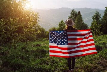 Patriot young caucasian woman in hat standing on background summer mountains during beautiful amazing sunset or sunrise, holding proudly usa flag high in air, demonstrates patriotism and freedom