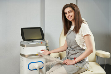 a beautiful smiling woman, a hair removal master, sits at a laser hair removal machine, holds a soft feather in her hands. depilation