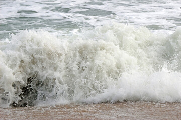 Close-up detail of white spray of a large ocean wave breaking in the shallows onto beach sand. No people.