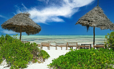 Fototapeta na wymiar Beautyful empty idyllic lonely bright white sand beach, 2 thatch umbrellas, row of wooden basic sun beds, turquoise water, clear blue sky - Paje, Zanzibar