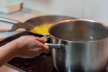 A woman's hand holding a pot that is boiling on a ceramic hob, preparing food.