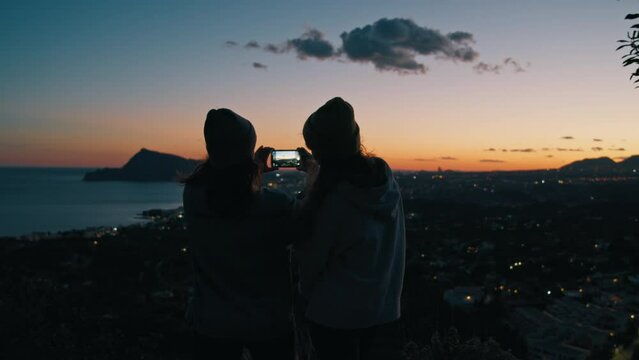 Young women, millennial girls, friends silhouette take colorful beautiful dark sunset seashore panoramic photo holding phone together, using camera on smartphone during weekend, trip, travel, vacation