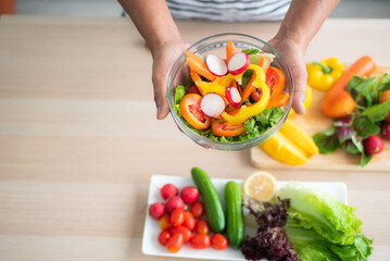 close-up top view of a vegetable salad holding in hand against a blurred background of vegetables on the table such as tomatoes, cucumbers, green oak, red oak, lemon in the kitchen.