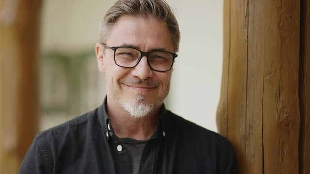 Portrait of happy casual middle age man in glasses standing on balcony at home relaxing, smiling.