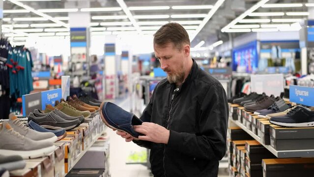 Man shopper chooses his shoes in a store, checking quality and thinking about buying. Slow motion. Inscription on shelf with shoes label in russian GENUINE LEATHER