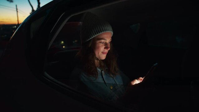Side night cinematic view of young beautiful woman sit in taxi car back seat near window on road trip, hold, check on mobile phone, use smartphone, scroll social media news, read messages and smile