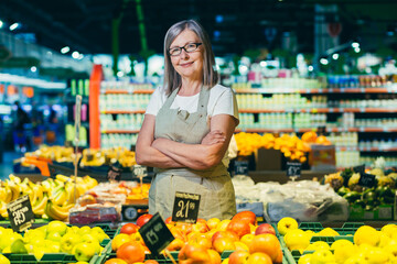 portrait of a senior gray-haired woman of a supermarket worker, worker spreads fruit with crossed arms smiles and looks at camera