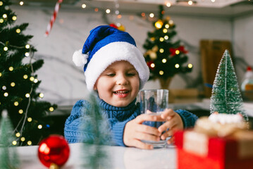 Boy toddler in a blue Santa hat drinking filtered water from a glass in the kitchen. Holidays, health concept.
