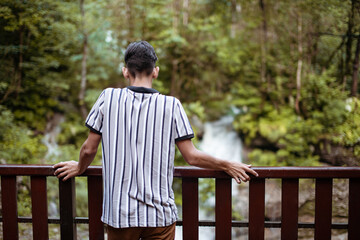 A young student stands with his back to the camera against the backdrop of a mountain river. Hiking in the mountains.