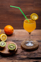 Orange fruit cocktail with a straw garnished with a slice of lemon in a large glass on a wooden table next to oranges and kiwi.