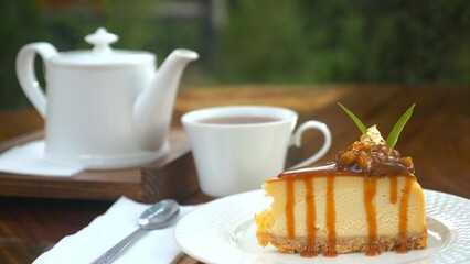 Piece of delicious fresh cheesecake cake on wooden table with white teapot in outdoor cafe. Tasty fresh pastry decorated with edible gold and caramel with cup of hot tea. English tea time. Sweet food