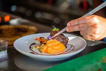 Close up of chef hands in hotel or restaurant kitchen decorating dish. Preparing beef steak with...