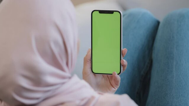 Rear view unknown islamic female muslim woman young lady girl in hijab lying on cozy sofa at home hold cellphone with green mobile screen display mockup for advertisement promotional image useful app