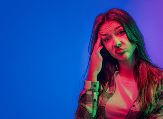 Portraits of young emotional woman on multicoloured background in neon lights. Looks doubtfully rubs his eyebrow with his hand. Place for text. Flyer Poster Banner