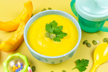 Pumpkin soup puree for the baby in a blue bowl, pumpkin seeds and pieces, a spoon, a drinking bowl and a toy on a yellow background. Baby nutrition, the first complementary food.