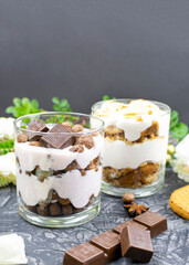Multi-layered cottage cheese dessert with chocolate corn balls, yogurt and milk chocolate slices in a glass on a black background. Romantic breakfast. Place for text, recipe.