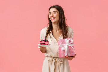 Celebration, party and happiness concept. Carefree smiling happy european woman having fun parying own b-day party, holding gift and piece cake, blow-out candle, pink background - obrazy, fototapety, plakaty