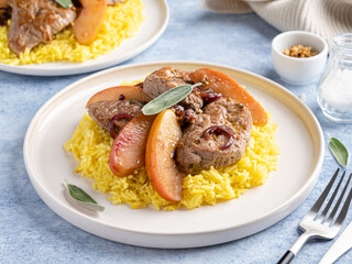 Pork chops stewed with apple slices, mustard and sage leaves served with curry rice. Food on white plate, fork and knife. Hot delicious dinner or lunch. Blue background. Close up view.