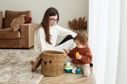 Mom and baby toddler pull toys out of basket. Babysitter and kid playing in room on floor.