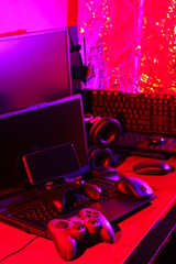Gamer's workspace. Gamepads, computer mouse, headphones, keyboard and laptop on the table. Gaming accessories and computer equipment in neon light. Gambling addiction concept