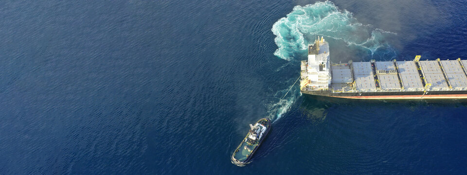Aerial drone ultra wide top down photo of tow - tug boat assisting by pulling empty container ship to depart from container terminal port