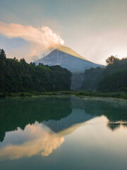Lake with a water surface that looks greenish and emits smoke in the morning with a volcano on the background in sunrise. The lake surrounded by trees. The volcano named Merapi Volcano in Indonesia.