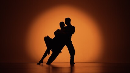 Couple of dancers approach each other and begin to dance Argentine tango. Elements of latin ballroom dance in studio with orange brown background. Dark silhouettes. Slow motion ready, 4K at 59.94fps. - obrazy, fototapety, plakaty