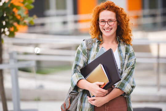 Beautiful Smiling Red Hair Student Girl Hugging Books Posing With Backpack Near College Building Outdoor. Modern Education, College Tuition And Grants, Studentship And Study Abroad Concept