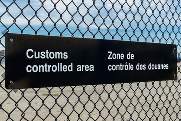 A bilingual Customs Controlled Area sign in English and French on a chain link fence. Angled view...