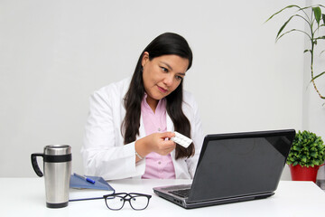 Latin woman doctor with white coat has online consultation by video call on the computer shows the pills that the patient has to take
