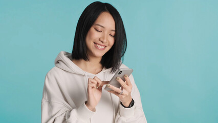 Pretty Asian woman wearing white hoodie chatting with boyfriends looking happy isolated on blue background. Female holding smartphone