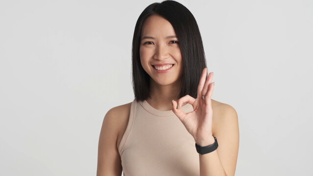 Confident Asian girl showing okay gesture smiling at camera over white background. Asian woman posing isolated. Like it