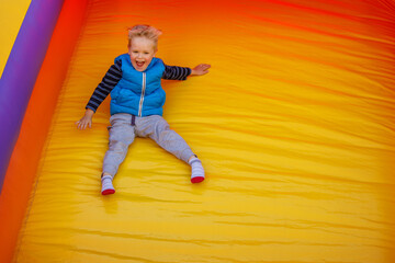 Happy little boy sliding down an a yellow- orange inflatable slide. There is free space for text in...