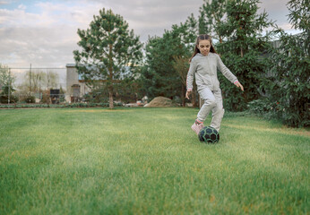 Cute little girl playing football with soccer ball on green lawn in backyard of house. Child kicking soccer ball on field. High quality photo