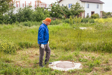 Man working plumber with a tire iron at a sewer well in the countryside. Maintenance of septic tanks and water wells - obrazy, fototapety, plakaty
