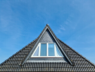 Roof window in velux style with black roof tiles against a blue sky.