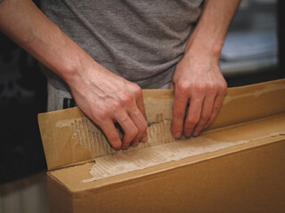 The hands of a caucasian young man in a gray tank top with sleeves open a large cardboard box