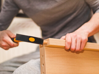 The hands of a Caucasian young man hammer a wooden nail into the headboard with a yellow hammer