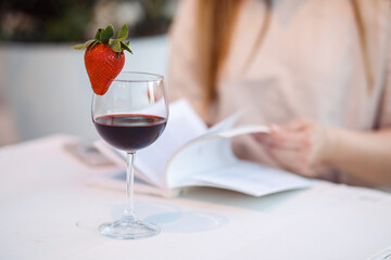 Close-up of wineglass with strawberry, cropped woman hands read book. Addiction to alcohol beverages. Celebration event