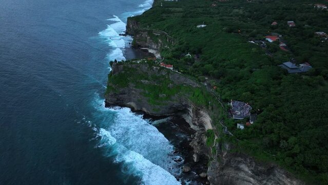 Aerial drone footage of Pura Luhur Uluwatu on sunset, one of Bali's most famous sea temples, perched on dramatic limestone cliffs overlooking the Indian Ocean. Waves crash against rugged coastline.