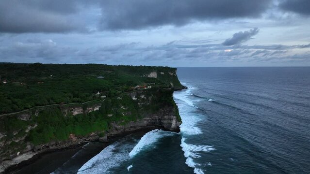 Aerial drone footage of Pura Luhur Uluwatu on sunset, one of Bali's most famous sea temples, perched on dramatic limestone cliffs overlooking the Indian Ocean. Waves crash against rugged coastline.