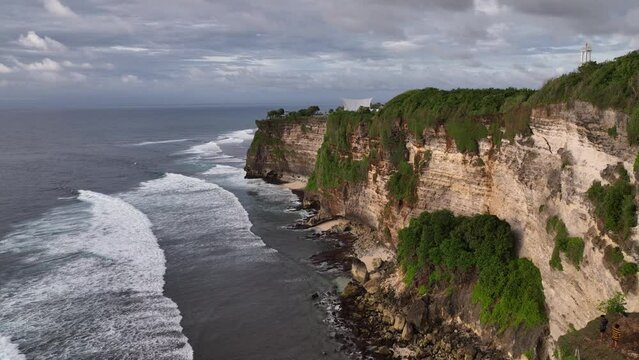 Aerial drone footage of Pura Luhur Uluwatu on sunset, one of Bali's most famous sea temples, perched on dramatic limestone cliffs overlooking the Indian Ocean. Waves crash against rugged coastline.