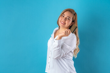 Beautiful attractive young business woman posing happily while holding credit card, in white shirt and grey pants. Isolated over blue background.