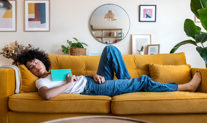 Tired African American woman sleeping on yellow couch after reading a book Female relaxing at home. - Powered by Adobe