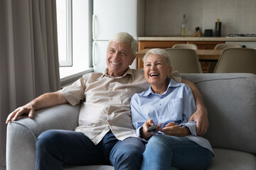 Laughing elderly spouses grey haired husband wife cuddle on couch hold remote controller watch comedy show movie on tv set. Overjoyed retired family ciuple spend cozy evening at home enjoy funny video