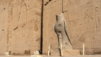 Granite sculpture of a falcon on the background of the stone wall of the temple of Horus in Edfu. Carvings of gods and hieroglyphs are visible. Egypt