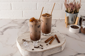 two tall glass with a chocolate drink or cold coffee with milk and ice cubes stand on a wooden podium opposite a white brick wall.