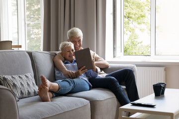 Modern tech for all ages. Bonding spouses retirees cuddle on couch at modern living room networking...
