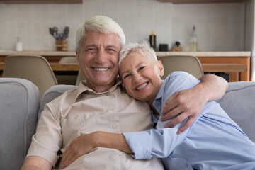 Headshot family portrait of two happy pensioners hoary senior adult husband wife cuddling on sofa. Loving family couple of grandparents laughing hugging looking at camera with healthy bright smiles