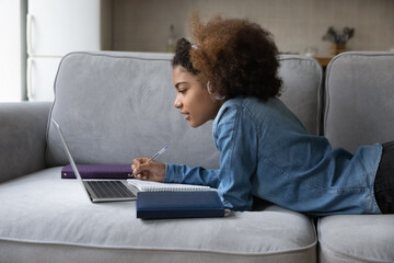 Focused Black high school student girl in headphones studying at home, lying on sofa at computer, watching learning webinar, lesson, video tutorial on laptop, writing notes for essay, class report